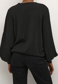 Person, der einen schwarzen Pullover mit breiten, gerippten Ballonärmeln und schwarze Jeans trägt, steht mit dem Rücken zu einer schlichten Wand.