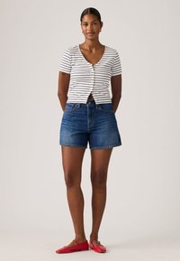 Γυναίκα που στέκεται φορώντας μια λευκή και μαύρη ριγέ μπλούζα με κοντά μανίκια, μπλε τζιν σορτς και κόκκινα φλατ παπούτσια σε ένα ουδέτερο φόντο.