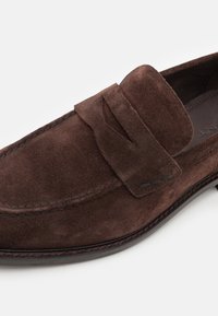 Brune semskede loafers med et stilrent design, der har en hævet remdetalje og syninger. Sålen er sort med en let kurve.