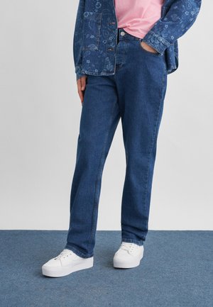 Persona che indossa jeans di denim blu, scarpe da ginnastica bianche, una camicia rosa e una giacca di denim blu con motivo floreale, con una mano in tasca.