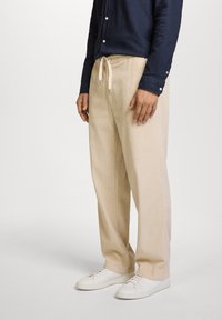 Pantalones de terciopelo beige con un patrón de estrías verticales y texturizadas, que cuentan con una cinturilla elástica con cordón, combinados con zapatillas blancas.