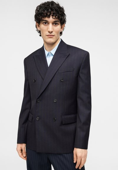Σκούρο μπλε διπλόσαρκο σακάκι με διακριτικές ρίγες, πέτο με κόψιμο, τσέπες με πατ πρόσθετες, και μαύρα κουμπιά, συνδυασμένο με ανοιχτό μπλε πουκάμισο.