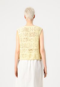 Keltainen neulottu lyhyt toppi, jossa hihaton muotoilu, pitsikuvio ja ruudukkomainen tekstuuri. Takaa näkyy avoin pitsityö.