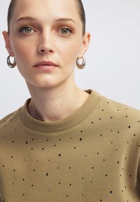 Khaki bluza z okrągłym dekoltem, ozdobiona rozsianymi czarnymi i srebrnymi ćwiekami. Gładka tekstura tkaniny; modelka nosi srebrne kolczyki w kształcie kół.