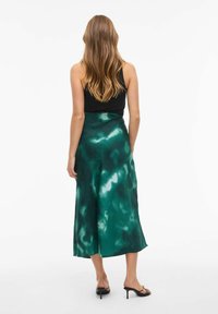 Jupe midi en soie verte avec un motif dégradé fluide ; texture lisse, ourlet large et fente ; associée à un débardeur noir et des chaussures minimalistes.