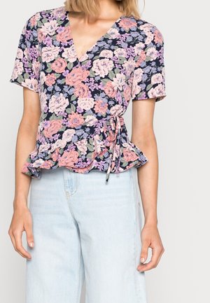 Femme portant un haut cache-cœur à manches courtes à motif floral avec ourlet volanté et un jean bleu clair taille haute, debout devant un fond uni.