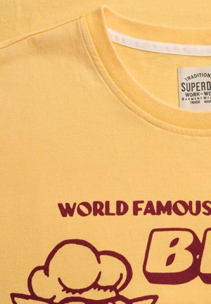 Żółta koszulka z okrągłym dekoltem, z bordowym napisem „WORLD FAMOUS” oraz fragmentem grafiki poniżej tekstu.