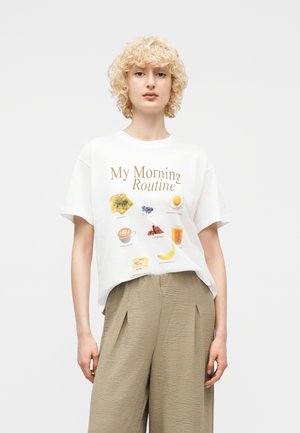 Jeune personne aux cheveux blonds bouclés portant un T-shirt blanc affichant des articles de petit-déjeuner et un pantalon beige à jambes larges, sur un fond uni.