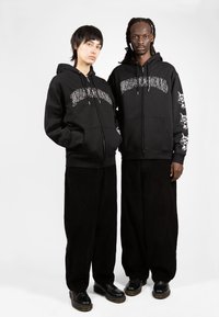 Μαύρες φούτερ με κουκούλα και λευκά γραφικά κείμενα και σχέδια στα μανίκια, φοριούνται με μαύρους φαρδιούς παντελόνες και μαύρα παπούτσια. Λείο υφασμάτινο υλικό.