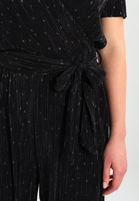 Svart plisserad jumpsuit med metalliska prickar, en insvept design och knytning i midjan, korta ärmar och en texturerad yta.