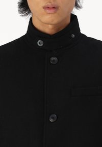 Abrigo de lana negro con cuello alto, que presenta un cierre de doble botón y un bolsillo en el pecho, textura suave, diseño minimalista.