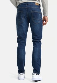 Jeans de un azul oscuro desgastado con un corte recto, cinco bolsillos y una etiqueta de cuero marrón en la cintura; combinados con zapatillas blancas.
