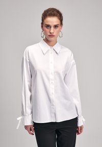 Camicia bianca a maniche lunghe con colletto a punta, tessuto liscio e polsini legati. Abbinata a pantaloni neri. Dettagli delicati sui polsini.