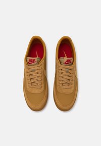 Nike tenisice od smeđeg kože s ukrasnim šavovima, ravnim vezicama i crvenom unutrašnjošću s Nike logotipom. Zaobljen vrh i teksturirana potplat.