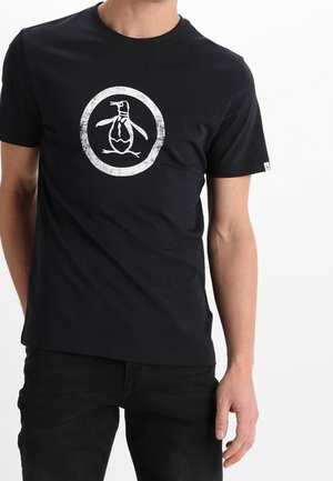 Homme portant un t-shirt noir avec un logo circulaire blanc représentant un manchot stylisé au centre à l'avant.