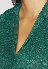 Grön texturerad tyg med metalltråd, med en sjalkrag och en v-ringad design. Närbild som framhäver lyster och ytdetaljer.