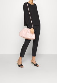 Rosa, mjukt texturerad axelväska med en samlad design och tunn rem. Bärs med svart topp och jeans, på en modell som står mot en neutral bakgrund.