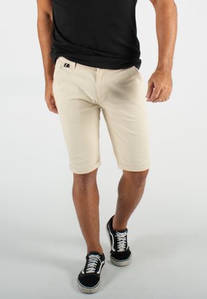 Shorts beiges à coupe slim, avec un ourlet roulé et des poches avant. Portés avec des sneakers noires à lacets et semelles blanches.