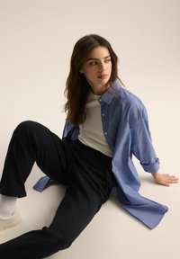 Niebiesko-biała w paski oversize'owa koszula z guzikami, zestawiona z czarnymi spodniami, na jasnym tle. Gładkie tekstury i swobodny krój.