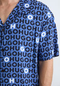 Short-sleeve shirt in blue fabric featuring a repetitive black and white text pattern. Buttons are visible on the front, accentuating the design.