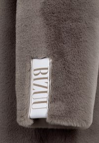 Grijze nepbont stof met een zachte textuur. Een aangehecht wit label toont "BIZUU" in beige hoofdletters.