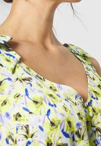 Nahaufnahme des Nackens und der Schulter einer Frau, die ein blumiges Kleid mit gelben, blauen und lila Blumen sowie mit Stoff überzogenen Knöpfen trägt.