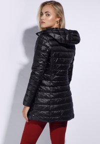 Zwarte gewatteerde jas met een capuchon, met een glanzende afwerking, gestikte textuur en een getailleerde snit, gedragen over rode broek.