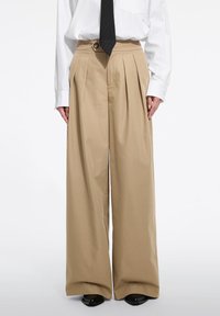 Pantaloni bej cu picioare largi, cu pliuri frontale, talie înaltă și un accent cu nasture. Asortați cu o cămașă albă și o cravată cu dungi negre.