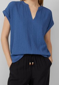 Top corto blu in tessuto strutturato con scollatura a V. Presenta un fit rilassato e maniche arrotolate, abbinato a pantaloni neri con coulisse e dettagli in perline.