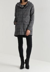 Mujer vistiendo un abrigo con capucha de cuadros grises y un cierre, una blusa negra con flores, pantalones negros ajustados y botines negros de tacón, de pie con una mano en el bolsillo.