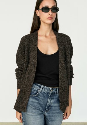 Frau trägt dunkelbraunen Strickcardigan, schwarzes Tanktop, blaue Jeans und schwarze rechteckige Sonnenbrille, steht vor einem schlichten hellen Hintergrund.