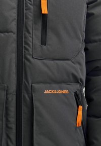 Giacca imbottita grigia con dettagli arancioni e tirazip. Presenta diverse tasche e il logo 'JACK & JONES' in arancione sulla parte frontale.