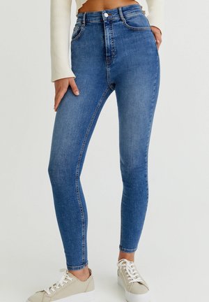 Kobieta w niebieskich obcisłych jeansach z wysokim stanem i beżowych trampkach, z jedną ręką opartą na udzie, a drugą w kieszeni.