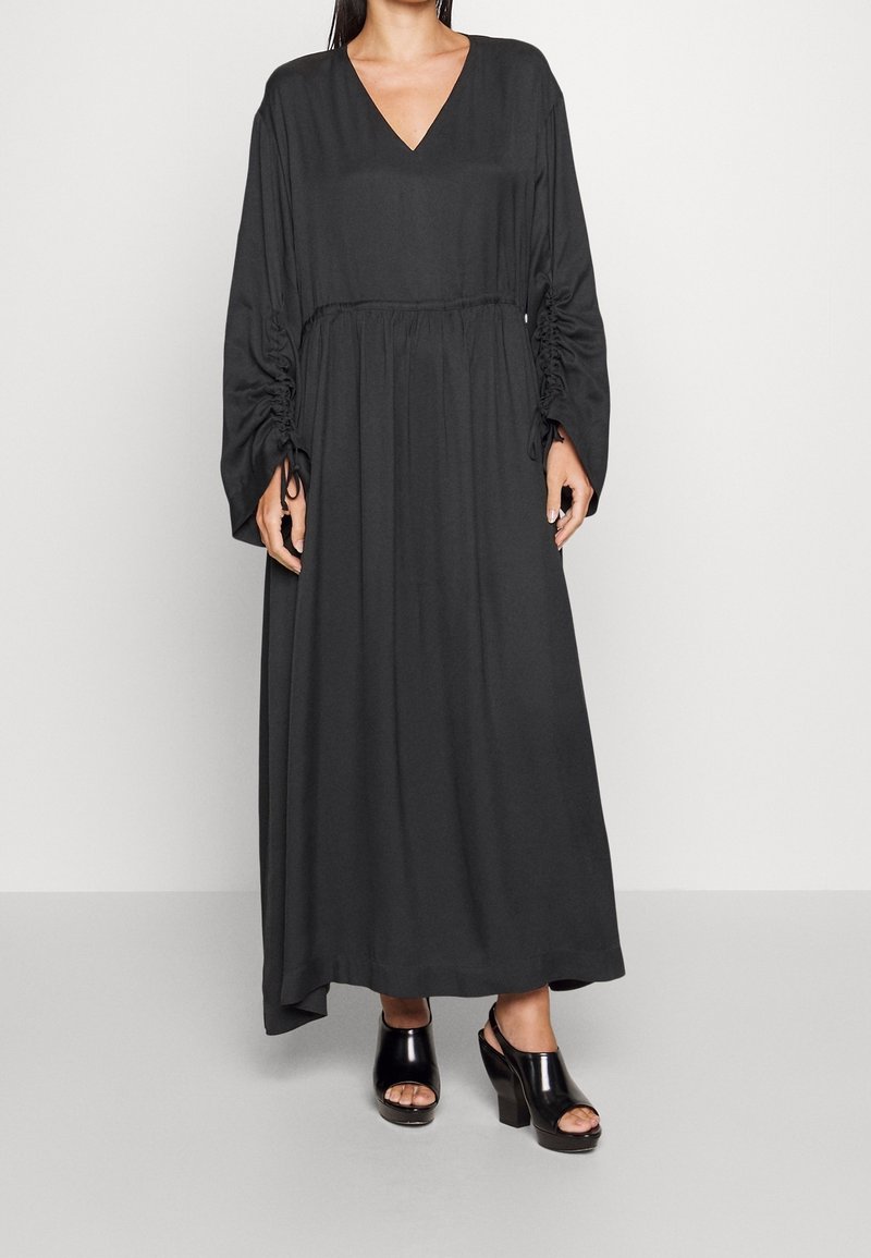 Schwarzes langärmeliges Kleid mit V-Ausschnitt und geraffter Taille, mit kräuseligem Akzent an den Ärmeln. Kombiniert mit schwarzen Plateauschuhen.