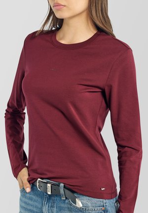 Mujer que lleva una camiseta ajustada de manga larga color granate con cuello redondo, vaqueros azul claro y un cinturón negro con hebilla plateada.