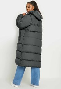 Abrigo de plumas largo y negro con capucha, con texture acolchada, bolsillos laterales y un corte holgado, llevado sobre jeans acampanados de un azul claro y zapatillas blancas.