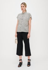 Chemise à manches courtes, boutonnée, avec un motif de pois noirs sur fond blanc ; assortie à un pantalon noir 7/8 et des sandales à talons noires.