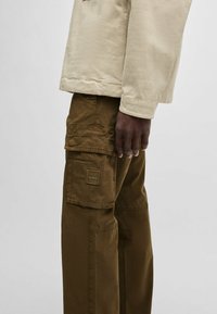 Beige jas met een relaxte pasvorm, gecombineerd met cargo-broeken in bruin. De broeken hebben meerdere zakken en een vierkant merklabel op de dij.