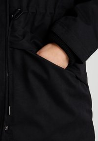 Zwarte buitenkleding met een gladde textuur, met een hand die in een zijzak rust en een manchetdetail van ribbelstof. Rits en knopen zichtbaar.