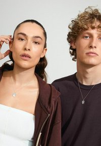 Silverhalsband bärs av två modeller, en i en vit topp och mörk hoodie; den andra i en mörk t-shirt. Naturliga hudtoner, enkla bakgrunder.