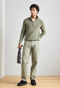 Oliven grøn ribbestrikket pullover med en kvart lynlås, en lysebeige t-shirt under, lyse olivengrønne cargo bukser og sorte slip-on sko. Sort skuldertaske.
