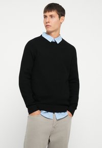 Black textured knit sweater with a round neckline, worn over a light blue and white striped collared shirt, paired with light gray pants.