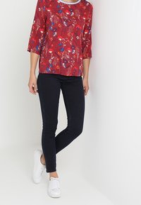 Top rosso con stampa floreale, maniche a tre quarti e scollatura rotonda, abbinato a jeans slim-fit scuri e sneakers bianche.