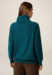 Türkiser, glitzernder Rollkragenpullover mit lockerer Passform und gerippten Bündchen, kombiniert mit braunen Hosen. Weiche Textur mit einem dezenten Schimmer.