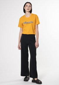 Gul bomulds t-shirt med grafisk tekst, parret med sorte brede bukser og sorte chunky sandaler, sat mod en ensfarvet hvid baggrund.
