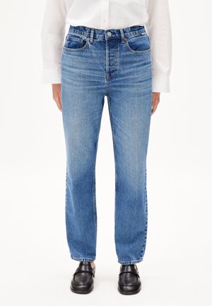 Person iført højtaljede blå jeans med lige ben, hvid skjorte med knapper og sorte lædersko uden snøring, stående på hvid baggrund.
