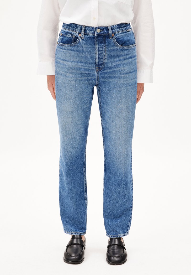 Personne portant un jean bleu taille haute à jambes droites, une chemise blanche boutonnée et des chaussures noires en cuir sans lacets, debout sur fond blanc.