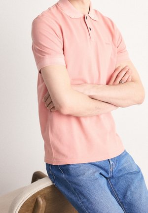 Person in einem pinken kurzärmeligen Poloshirt und blauen Jeans steht mit verschränkten Armen und lehnt an einem Holzstuhl mit beiger Polsterung.