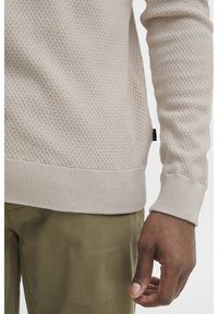 Ljusbeige texturerad tr�öja med ribbat nederkant och ärmslut, bärs över olivgröna byxor. Närbild på ärmen och en del av torso.