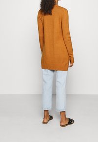 Hořčicově žlutý svetr s dlouhým rukávem a žebrovaným lemem, v kombinaci se světle modrými zkrácenými džíny a černými sandály, viděný zezadu.